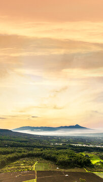 Aerial view of lush agricultural fields and rolling hills at sunset with a distant mountain range under a golden sky in Bumiaji, East Java, Indonesia.