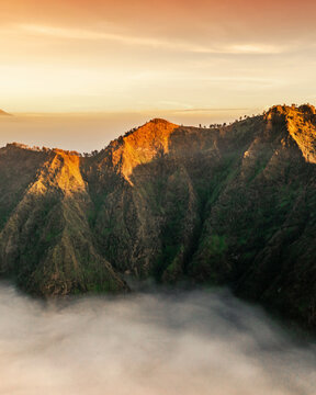 Aerial view of Mount Bromo at sunrise with golden light illuminating the rugged volcanic peaks rising above a sea of clouds in Tosari, East Java, Indonesia.
