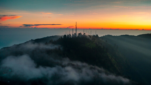 Aerial view of Mount Bromo area with telecommunication towers atop a misty, forested mountain peak during a vibrant sunset with golden light in Tosari, East Java, Indonesia.