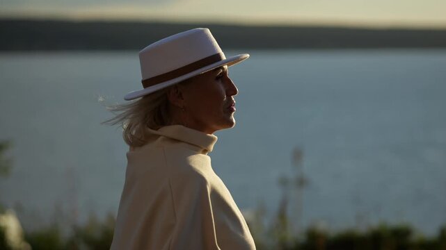 Woman moves past tranquil shoreline in warm evening light, dressed in pale coat and hat. Lakeside walk creates peaceful, stylish moment with soft wind and thoughtful expression beside calm water.