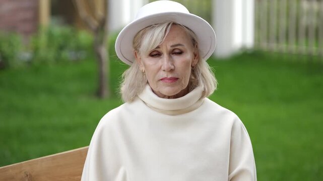 Sad elderly woman sits on bench in garden, eyes lowered and face tense. White hat and soft cream clothing heighten melancholy mood, while her quiet pose suggests deep sorrow and depression.