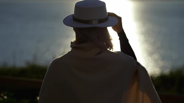 Backlit woman adjusts hat beside water. Woman stands in soft evening light, facing bright reflection while touching hat brim, creating calm portrait with elegant silhouette and warm glow.