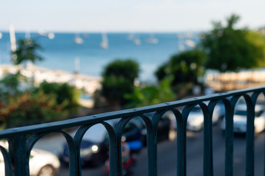 A dark metal balcony railing stands in sharp focus in Kerkira, Greece. A blurred coastal street and blue sea filled with boats extend into the background.