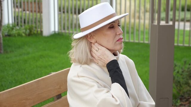 Seated woman presses neck with pain. Elderly woman in white hat and light shawl rests on bench outdoors, eyes closed and jaw tight, conveying discomfort beside trimmed grass and fence.