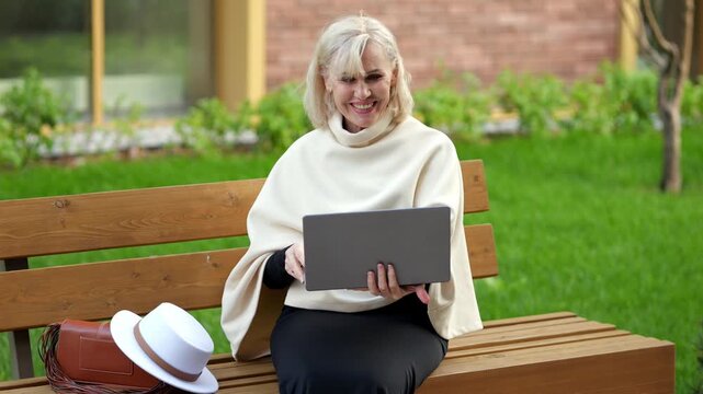 Woman using laptop on park bench takes part in online conversation. Smiling older woman waves at screen, then rests hand on chest while seated with handbag and white hat beside her.