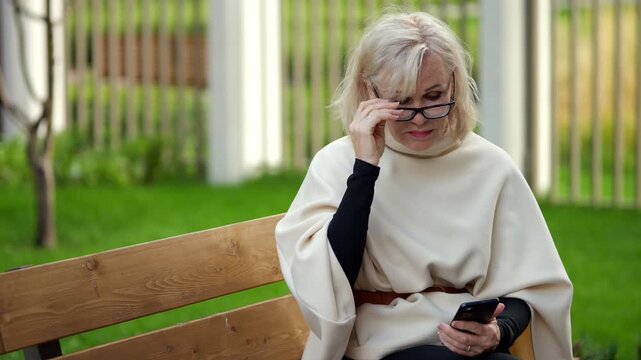Woman on park bench holds smartphone during outdoor break. She lifts eyeglasses, then settles them on her face while sitting beside green lawn, keeping calm, thoughtful mood in soft daylight.