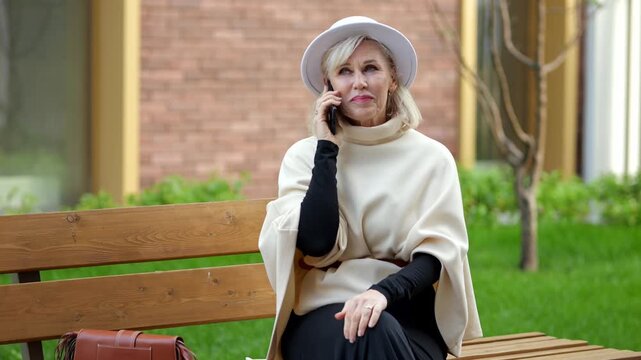 Woman on park bench takes smartphone call. She sits outdoors in courtyard, speaking with calm focus while holding phone to her ear and resting beside handbag on bench, listening during conversation.