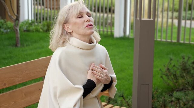 Older blonde woman rests hand over heart on park bench, showing pain and inward tension. Woman in soft cream clothing sits quietly in garden surroundings, breathing through strained moment.