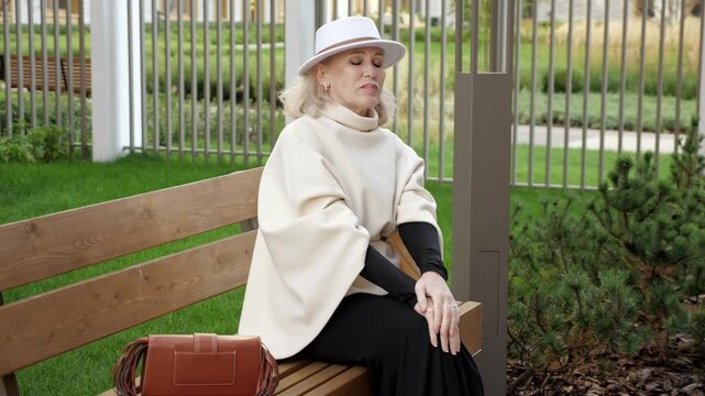 Knee pain troubles older woman seated on wooden bench, with hand on knee and tense expression. Her white hat and cream shawl add refined style to calm garden surroundings as she rests her leg.