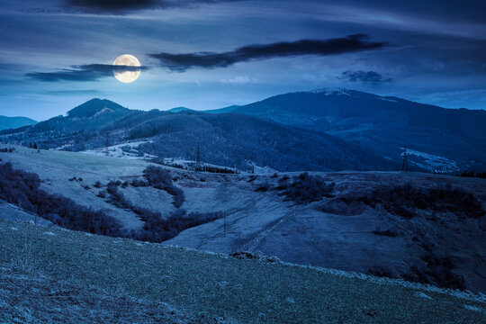 mountainous rural landscape of ukraine on spring night. picturesque highland under dark sky. grassy meadow in full moon light. carpathian countryside scenery with forest on a hill behind the pasture