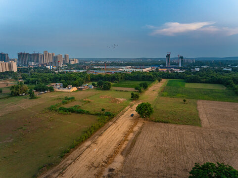 Aerial view of a dirt road cutting through agricultural fields towards the modern skyline of Gurgaon, Haryana, India.