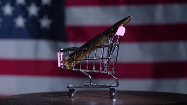 Close up of a bullet placed in a miniature shopping cart against USA flag background symbolizing gun trade consumerism and political debate concept, rotates