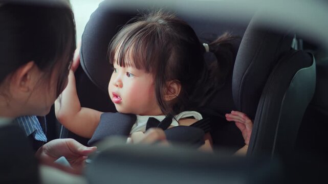 Caring Asian mother talking to toddler daughter while checking safety harness on child car seat, parent securing little girl before travel, family transportation, child protection