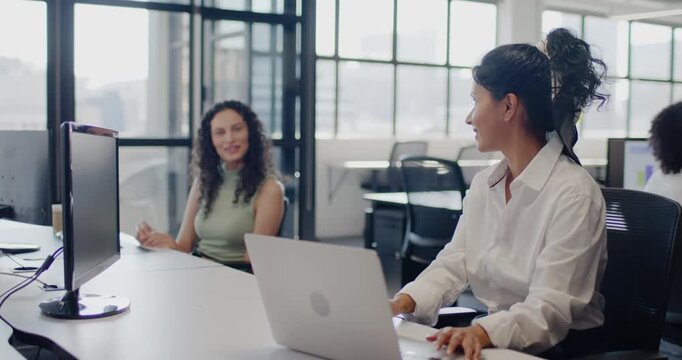 Diverse female team sharing info at desk by window, right woman pointing at laptop, left responding