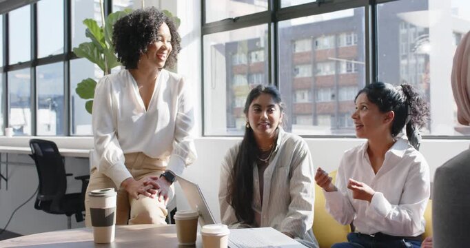 Diverse female coworkers in office, right woman starting discussion, gesturing at laptop, planning