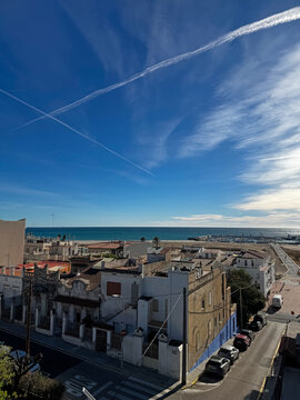 Scenic view of a coastal city by the sea in Spain, with a blue sky crossed by long white contrails left by passing airplanes.