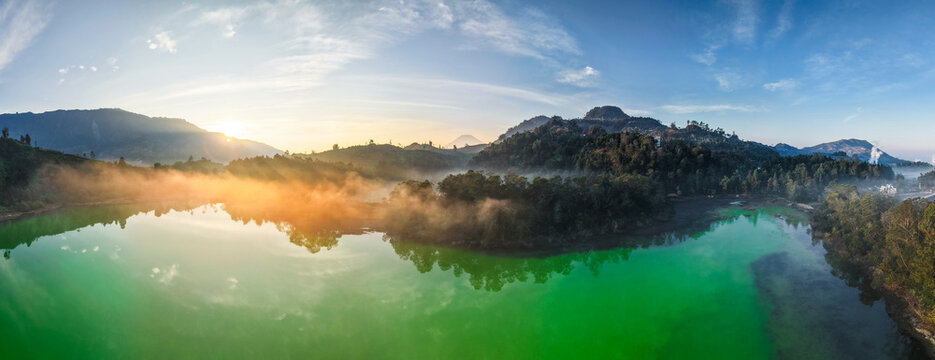 Aerial view of Telaga Warna Dieng with its vibrant green volcanic lake surrounded by misty hills and lush forest under a golden sunrise in Indonesia.