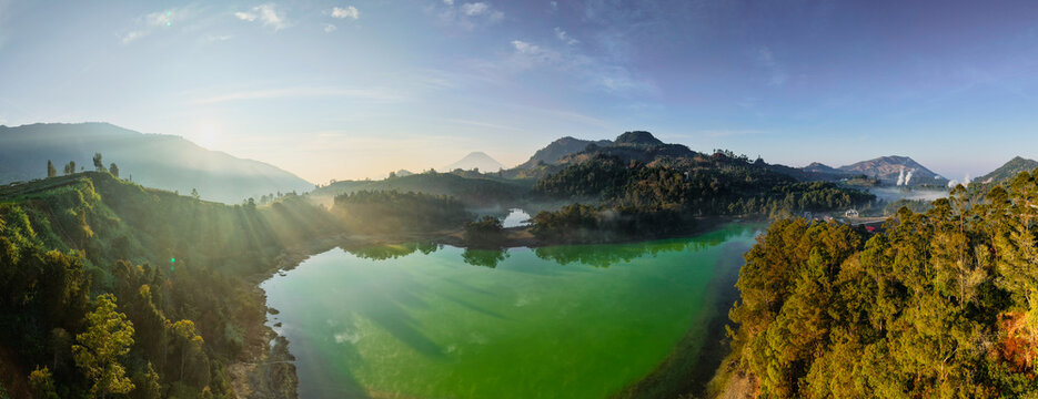 Aerial view of Telaga Warna Dieng with its vibrant green volcanic lake surrounded by lush forested hills and misty atmosphere under a bright morning sky in Indonesia.