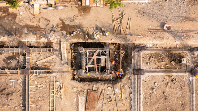 Aerial view of a construction site with workers in safety gear, steel reinforcement bars, wooden planks, and foundation pits in Klaten, Central Java, Indonesia.