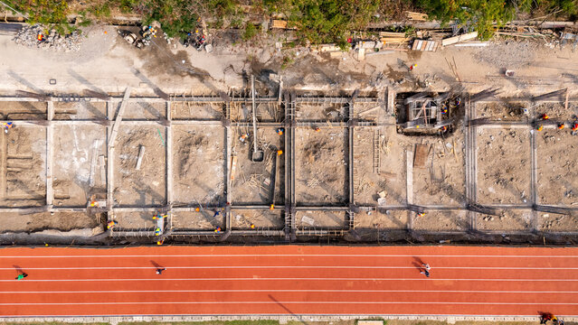 Aerial view of a construction site with building foundations and workers in orange vests next to a red running track in Klaten, Central Java, Indonesia.