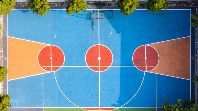 Aerial view of a vibrant, multi-colored basketball court with blue, orange, and red sections, surrounded by lush green trees in Klaten, Central Java, Indonesia.