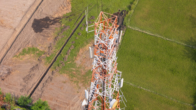 Aerial view of a telecommunications tower standing amidst vibrant green rice fields and brown tilled soil in Klaten, Central Java, Indonesia.
