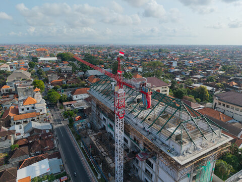 Aerial view of the construction site of the Wangaya Regional General Hospital featuring a prominent red and white tower crane and steel roof framework in Kecamatan Denpasar Utara, Bali, Indonesia.