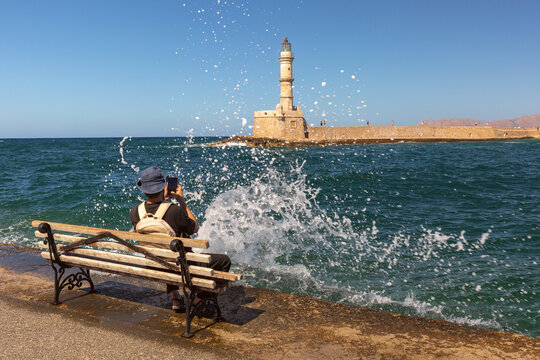 Tourist taking a photo of the lighthouse from the waterfront in Chania on Crete, Greece, with sea waves splashing against the shore.