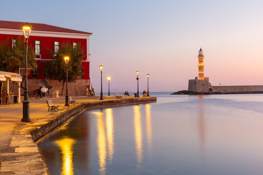 Illuminated lighthouse and waterfront promenade reflected in calm harbor water at sunset in Chania, Crete, Greece.