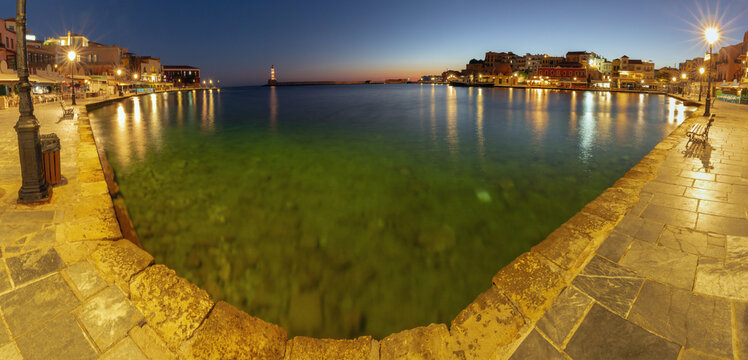 Panoramic view of Chania harbor at sunrise with illuminated waterfront buildings and reflections on the water, Crete, Greece.