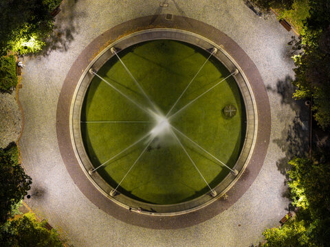 Aerial view of a circular fountain illuminated at night with water jets and surrounding stone pavement in a park in Warsaw, Poland.