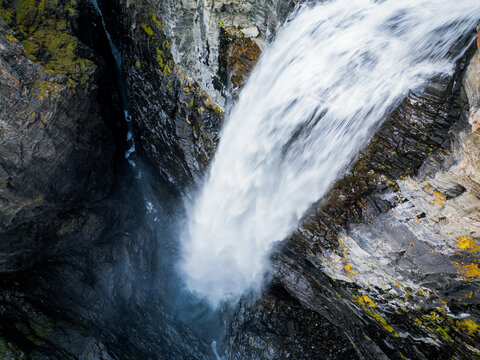 Aerial view of the powerful Gorsa waterfall cascading into a deep, narrow rocky canyon with dark, mossy cliff walls in Birtavarre, Troms, Norway.