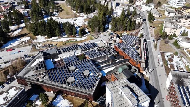 congress centre rooftop solar panel array covers timber clad building davos switzerland cars circle roundabout below dense townscape backs onto forested mountainside aerial drone footage overhead pass
