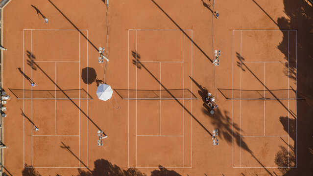 Aerial view of clay tennis courts with white lines and nets, featuring players and long shadows cast across the surface in Montevideo, Departamento de Montevideo, Uruguay.