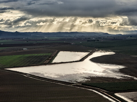 Aerial view of flooded agricultural fields under dramatic storm clouds with sunbeams breaking through in Andalusia, Spain.