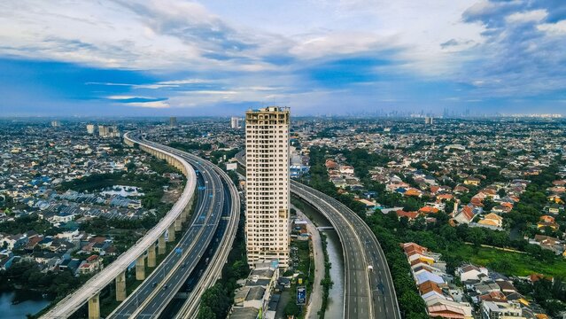 Aerial view of a tall residential building surrounded by a sprawling urban landscape with a multi-lane elevated highway winding through the city in Bekasi Barat, West Java, Indonesia.