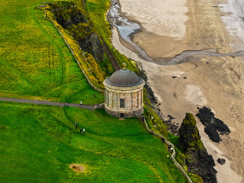 Aerial view of the historic Mussenden Temple perched on a grassy cliff edge overlooking the sandy beach of Downhill Strand with people in Coleraine, Causeway Coast and Gl, United Kingdom.