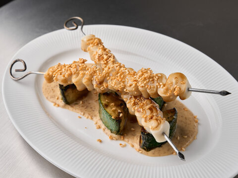 Sesame coated grilled vegetable skewers on white plate, creamy sauce pooling beneath, close up shot highlighting sesame seeds, char marks on zucchini, fresh herb garnish, appetizing presentation