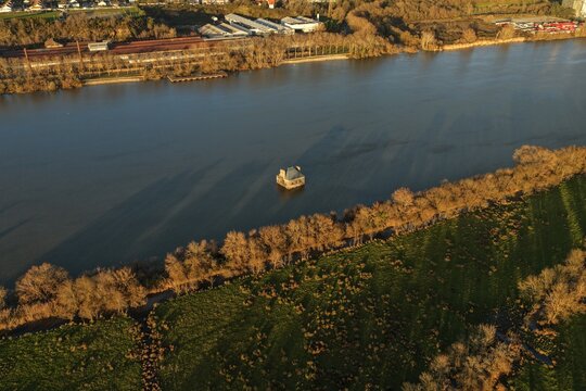 Coueron, France - 29 December 2025: Aerial view of La Maison dans la Loire, a house sculpture partially submerged in the river, surrounded by trees and fields in golden light.