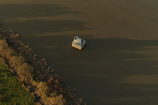 Coueron, France - 29 December 2025: Aerial view of La Maison dans la Loire, a surreal house sculpture partially submerged in the river, with calm water and grassy banks in golden light.