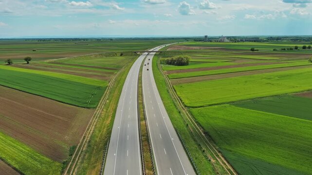 Aerial view of highway crossing green agricultural fields in Serbia. Cars driving on road through flat farmland plain under blue sky, 4k