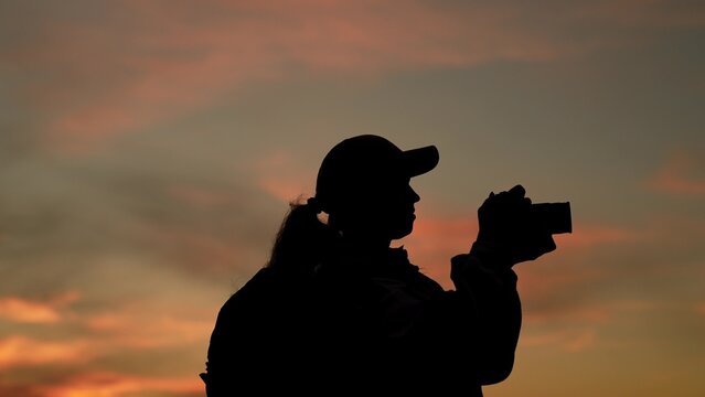 Female traveler with camera, backpack, silhouette. Photographer capturing beautiful nature shots at sunset. Female videographer filming stunning landscape at sunrise, magnificent sky. Woman tourist