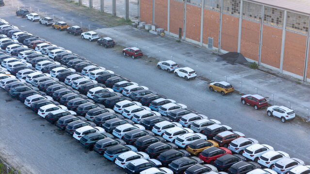 Piacenza, Italy - 01 August 2026: Aerial view of a car parking factory yard filled with rows of new vehicles parked on a gravel lot next to a large industrial brick building.