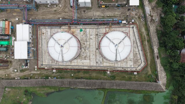 Aerial view of two large industrial storage tanks in a containment area with surrounding pipes and infrastructure near a pond and greenery
