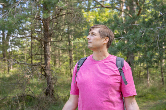 Caucasian man in his 40s wearing pink T-shirt with backpack looking away in pine forest in sunlight