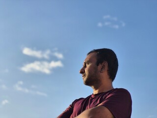 Side Profile Portrait of a man standing outdoors in the sun with Blue Sky and clouds in the background, Golden Hour, Peaceful and Calm, Crossed Arms, A Man Enjoying the Evening. Healthy Lifestyle. © Nwz