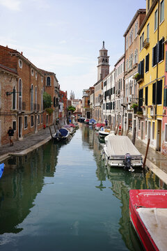 A tranquil Venetian canal lined with colorful buildings and moored boats, leading toward a distant bell tower on a sunny day.
