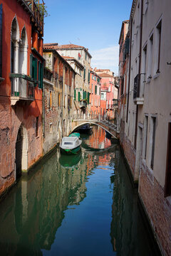 A stone arched bridge crosses a tranquil Venetian canal lined with terracotta buildings and a moored boat.