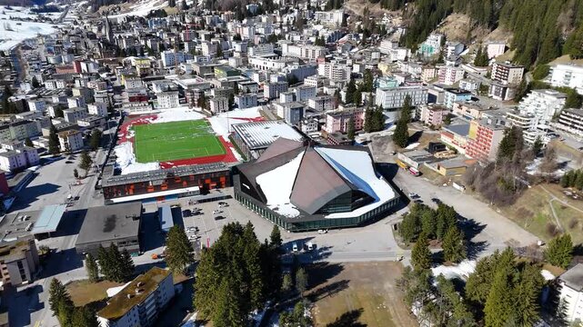 ice hockey arena angular roof panels glass frontage overlooks football stadium red track green pitch tourismus sport zentrum davos townscape forested mountainside snowcapped peaks switzerland