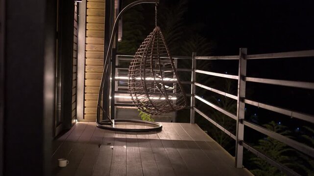 Rattan hanging swing chair gently swaying on a modern wooden balcony at night with warm exterior lighting, creating a tranquil and relaxing atmosphere in a cozy outdoor space